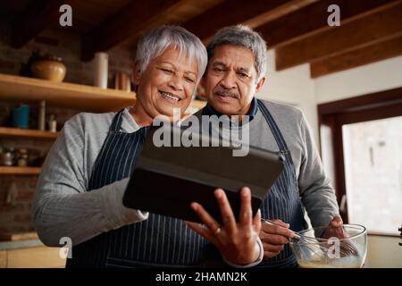 Multi-cultural elderly couple smiling, using tablet to research recipe in modern kitchen. Happy couple cooking healthy meal. Stock Photo