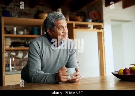 Pensive multi-cultural elderly male, leaning on kitchen counter with coffee mug in modern kitchen. Stock Photo