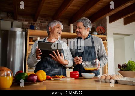 Multi-cultural elderly couple laughing together while cooking breakfast in modern kitchen, using tablet to research healthy recipe. Stock Photo