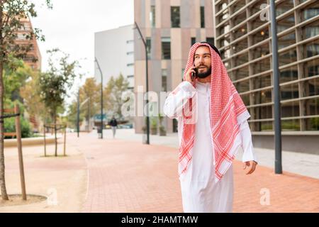 Arab businessman in traditional clothes making call on cellphone, talking on mobile phone with business partner while walking city street in Dubai, Saudi arab man using smartphone outdoors Stock Photo