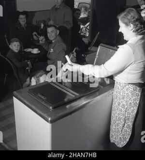 1950s, historical, inside a cafe, seated customers look on, as a little girl, having put some coins on the lid of the chest freezer, is given an ice cream wafer from the lady owner, London, England, UK. Stock Photo