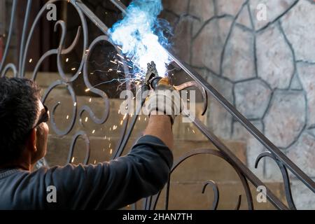 Man in safety glasses is welding metal of staircase. Welding work on metal in private house. Close-up. Stock Photo