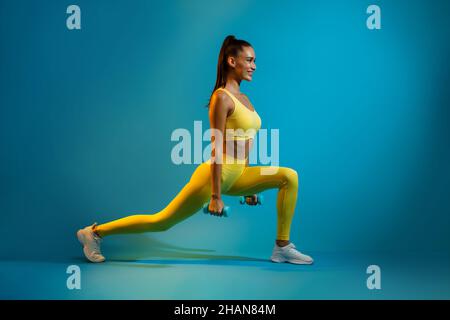 Sporty Female Doing Deep Lunge Exercise Holding Dumbbells, Blue Background Stock Photo