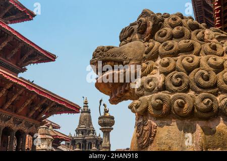 A stone guardian lion in front of the old royal palace in Durbar Square, Patan, Nepal.  Behind is the statue of King Yoganarendra Malla and the Vishmu Stock Photo