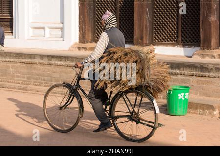 A Nepali man in a dhaka topi cap carries bundles of natural brooms on a bicycle in Durbar Square, Bhaktapur, Nepal. Stock Photo
