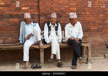 Three older Nepali men wearing traditional dhaka topi hats sit on a bench together in Durbar Square in Patan, Nepal.  One naps. Stock Photo