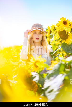 A blonde pretty long haired young women (22 years) in a sunflower field on a bright sunny day wearing a raffia straw hat editorial lifestyle image. Stock Photo