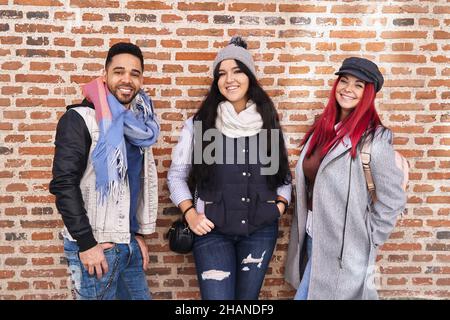 Positive multiracial young male and female friends wearing trendy casual outfits standing near brick wall and smiling at camera Stock Photo