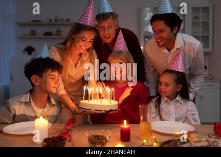 Happy multi generation family having birthday party, giving cake with lit candles to senior woman, congratulating her Stock Photo