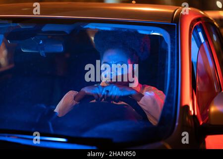 Depressed afro woman sit behind steering wheel in car at night, pondering about problems in life Stock Photo
