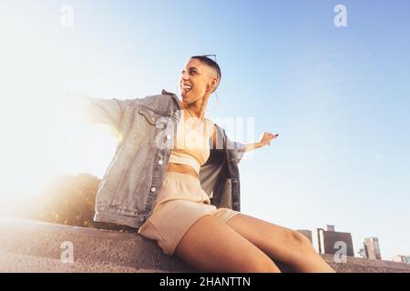 Young woman celebrating with her arms outstretched in the city. Happy young woman sitting on a wall outdoors in the sun. Cheerful young woman feeling Stock Photo