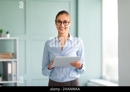 Positive business lady in formal wear using tablet in office, looking and smiling at camera, standing near workplace Stock Photo