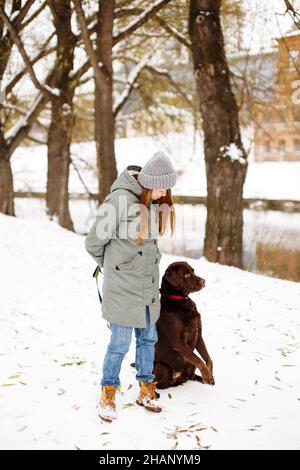 Smiling young woman with brown dog labrador retriever in winter warm outfit walking outdoors in snowy park, full body looking at camera Stock Photo