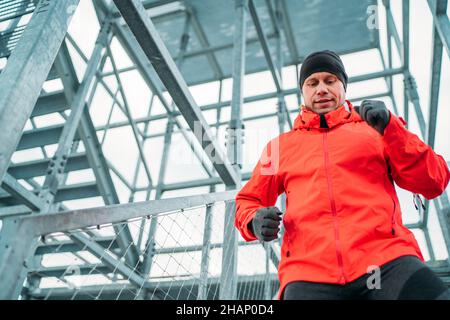 Waist portrait of smiling runner man dressed bright red softshell sporty clothes and gloves running down by huge steel industrial stairs in cold winte Stock Photo