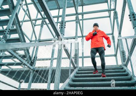 Smiling runner man dressed bright red softshell sporty clothes running down by huge steel industrial stairs in cold winter day. People healthy lifesty Stock Photo