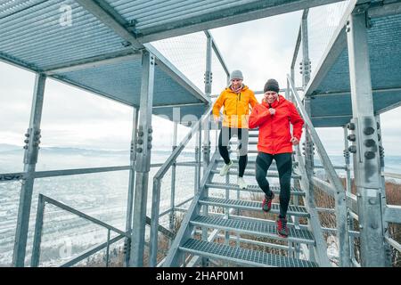 Two cheerfully smiling bright sporty clothes dressed men running down by huge steel industrial stairs with picturesque winter city landscape view. Men Stock Photo