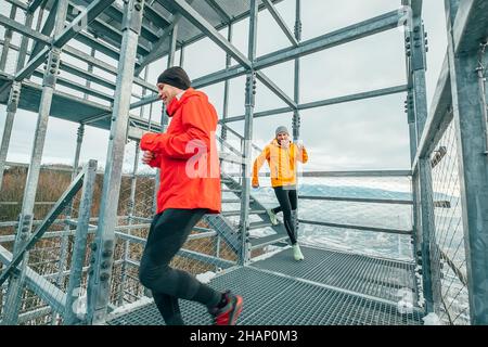 Two cheerfully smiling bright sporty clothes dressed men running down by huge steel industrial stairs with picturesque winter city landscape view. Men Stock Photo