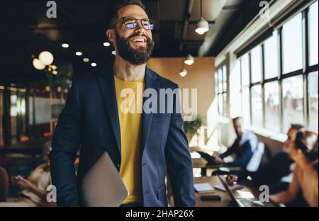 Happy young businessman smiling in a co-working space. Contemplative businessman looking away thoughtfully while holding a laptop. Cheerful young entr Stock Photo