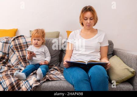 A woman is sitting on the sofa and reading a book. A small child is sitting next to him and playing on the phone. Family relax. Stock Photo