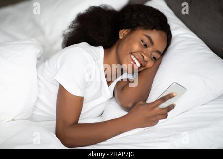 Positive young black lady laying in bed with smartphone Stock Photo