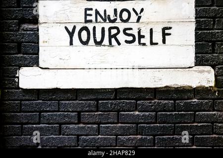Words on the wall - Enjoy Yourself on a boarded over window surrounded by bricks painted black. Stock Photo