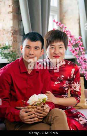 Happy young Asian couple in smart casualwear sitting on couch in front of camera in home environment Stock Photo