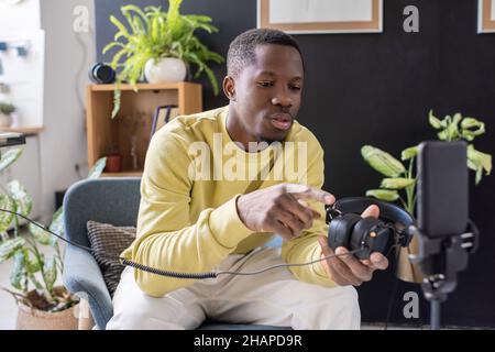 Young African man with headphones in hand giving masterclass or livestream in front of smartphone camera Stock Photo