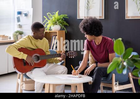 Teacher of music playing guitar in front of his student sitting by table and making notes at lesson in home environment Stock Photo
