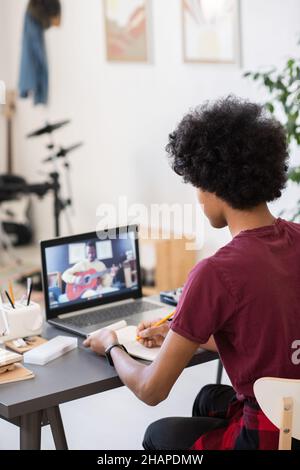 Biracial guy making notes while sitting by table and looking at music teacher with guitar on laptop screen Stock Photo