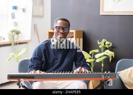 Happy young man in eyeglasses and casualwear sitting by piano keyboard and looking at camera in home environment Stock Photo