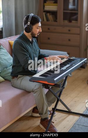 Side view of young teacher of music in headphones sitting on couch by piano keyboard and pressing keys Stock Photo