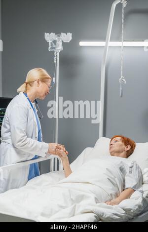 Mature female doctor holding hand of her patient and talking to her while she lying on hospital bed Stock Photo