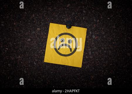 A sticky note with a sad face on it pinned to a corkboard. Close up. Stock Photo