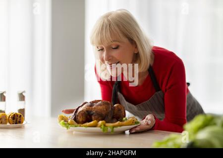 Beautiful senior woman smelling tasty roasted turkey, cooking festive Christmas dinner for her family in kitchen, copy space Stock Photo