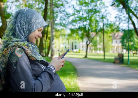 Profile view of the muslim pregnant woman smiling while using mobile phone and sitting alone in the park Stock Photo