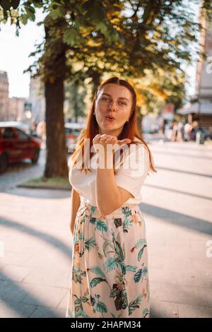 Portrait of young beautiful smiling 30s female in blowing kiss during summer time. Carefree positive woman posing, having fun on city street backgroun Stock Photo
