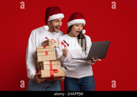Online Sales. Surprised couple wearing santa hats using laptop and credit card Stock Photo
