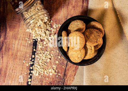 Overhead view of homemade honey raisin oatmeal cookies in a black bowl and fallen oatmeal flakes on the wooden table. Concept of natural and healthy e Stock Photo