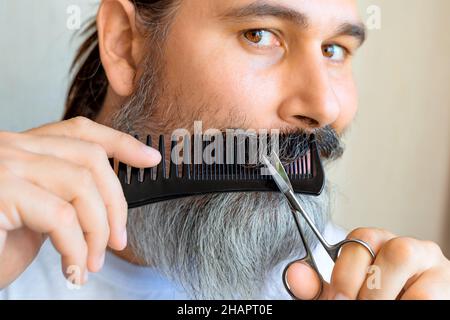 Middle-aged caucasian bearded man cut his mustache. A gray-bearded man cuts his own mustache using a comb and scissors. Selective focus. Stock Photo