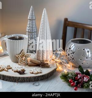 A cup of coffee on a wooden stand, ceramic Christmas trees in pastel colors, gingerbread cookies, gray candlestick. Artificial spruce branch with red Stock Photo