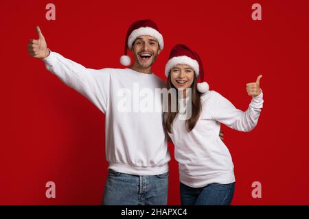 Christmas Offer. Cheerful Young Couple In Santa Hats Gesturing Thumbs Up Stock Photo