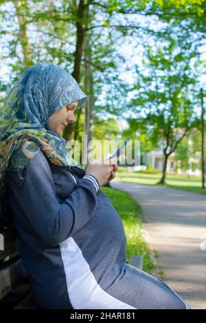 Profile view of the muslim pregnant woman using mobile phone and sitting alone in the park Stock Photo