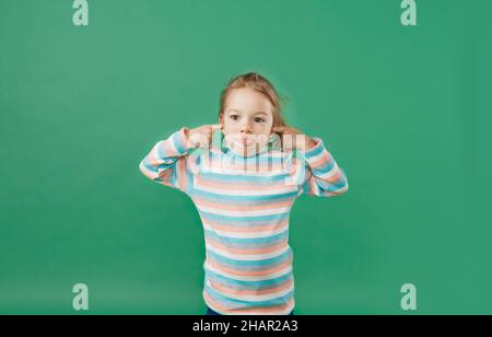 a little girl in a striped sweatshirt makes a face and shows her tongue on an isolated green background Stock Photo