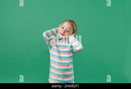 a little girl in a striped jacket covers her ears with her hands on an isolated green background Stock Photo