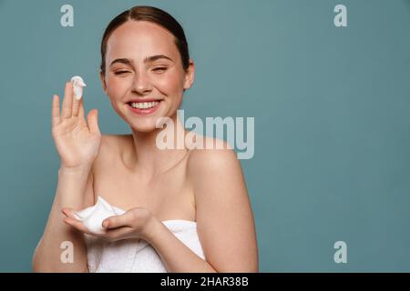 Ginger woman wearing towel smiling while showing cosmetic foam isolated over blue background Stock Photo