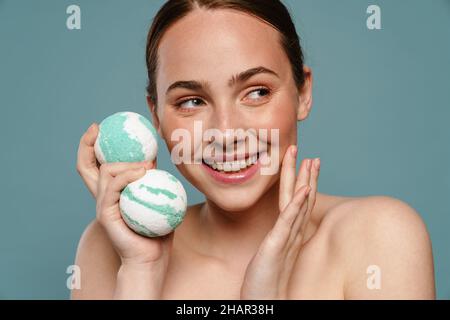 Ginger woman wearing towel smiling while showing balls bath bomb isolated over blue background Stock Photo