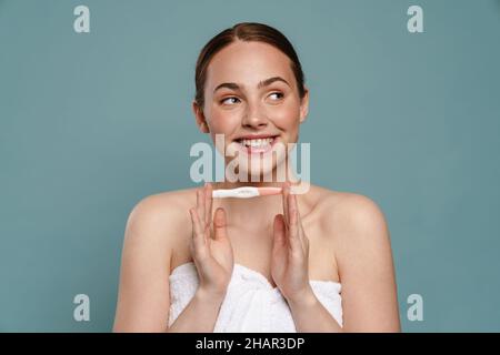 Ginger woman wearing towel smiling and showing pregnancy test isolated over blue background Stock Photo