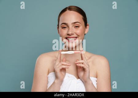 Ginger woman wearing towel smiling and showing pregnancy test isolated over blue background Stock Photo