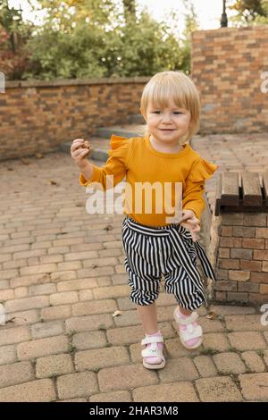 A beautiful cheerful little girl in a stylish striped suit plays with autumn leaves in a city park. Lifestyle Stock Photo