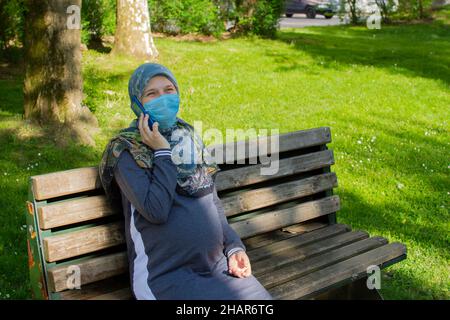 Young muslim pregnant woman wearing face mask talking on the phone while sitting in the park Stock Photo